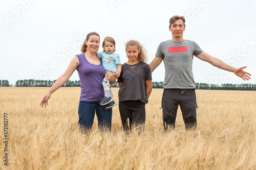 Husband and wife with their preteen age daughter and toddler child, mother holding baby on arms, four Caucasian people family standing on golden wheat field