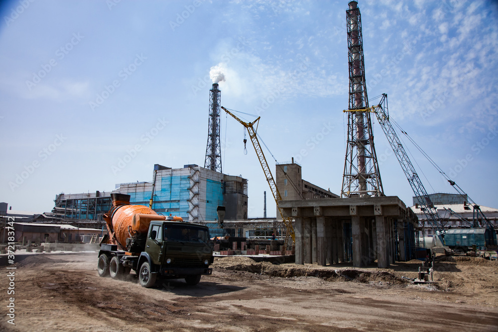Concrete construction, yellow mobile cranes, orange concrete mixer truck, blue factory building and smoke stacks on blue sky background. Renovation on chemical plant of phosphoric fertilizers.