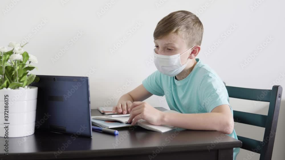 A young boy in a face mask talks to someone on a tablet as he does homework for school at a table at home A young boy in a face mask talks to someone on a tablet as he does homework for school at a table at home