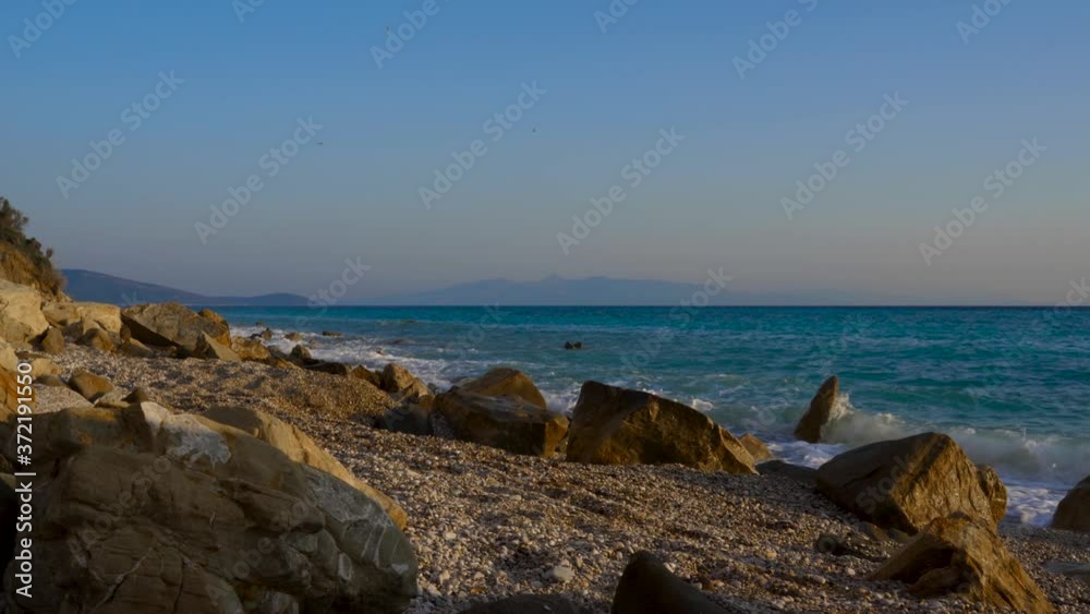 Sea waves splashing on rocks of peaceful beach with pebbles, seascape with Corfu island in background Sea waves splashing on rocks of peaceful beach with pebbles, seascape with Corfu island in background