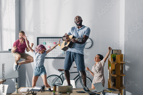 Selective focus of american businessman playing acoustic guitar while multiethnic colleagues dancing in office