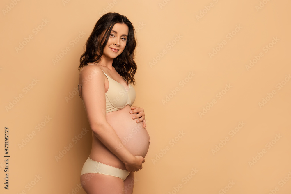 brunette pregnant woman in underwear touching belly and looking at camera on beige brunette pregnant woman in underwear touching belly and looking at camera on beige