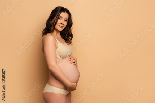 brunette pregnant woman in underwear touching belly and looking at camera on beige