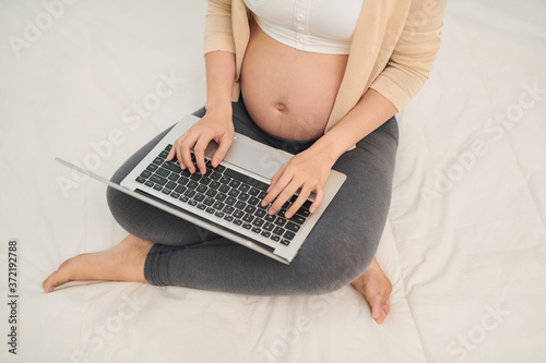Pregnant woman in white clothes sitting and using laptop in bed.