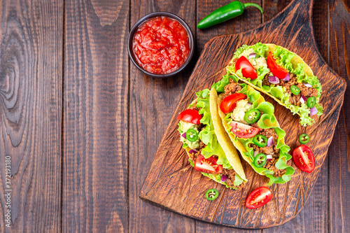 Taco shells on wooden board, with lettuce, ground beef meat,  mashed avocado, tomato, red onion and jalapeno pepper, horizontal, top view, copy space