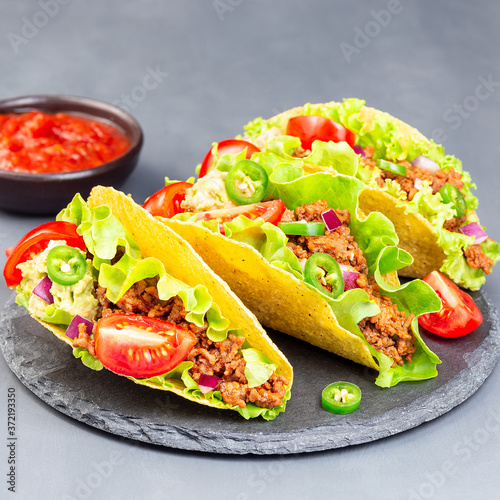 Taco shells with lettuce, ground beef meat,  mashed avocado, tomato, red onion and jalapeno pepper, on stone plate, square