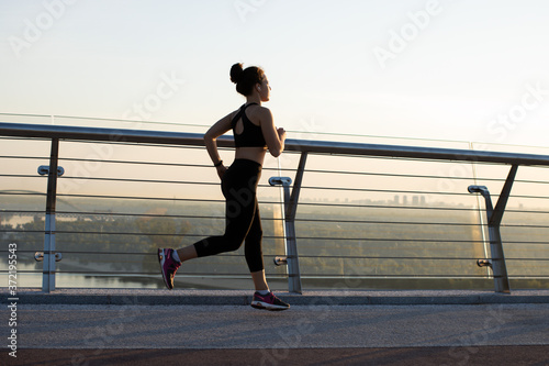 Running woman. Young fit smiling female runner in wireless headphones jogging during outdoor workout on the bridge in the morning sunrise. Sport healthy and active lifestyle concept © Parten