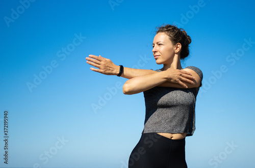 Fit and sporty young brunette woman stretching arms during exercising outdoors before running,standing on blue sky background in sunny summer day.Prepare muscles and ligaments before training workout