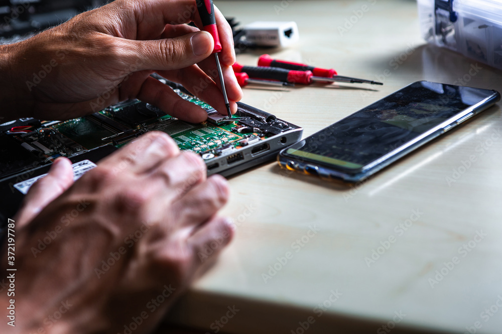 Fototapeta premium The service technician is trying to repair the laptop computer that is on his desk. Around the computer is a tool and a mobile phone on which he looks at how to do it.
