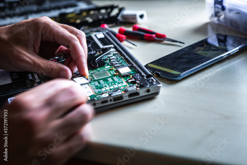 The service technician is trying to repair the laptop computer that is on his desk. Around the computer is a tool and a mobile phone on which he looks at how to do it.