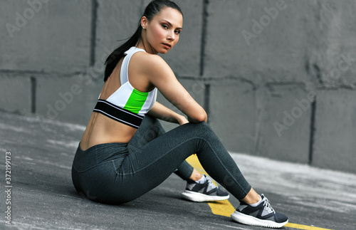 Determined athlete young woman sitting next to the gray concrete wall after workout outdoor. Fitness girl resting after exercising in the morning outside.
