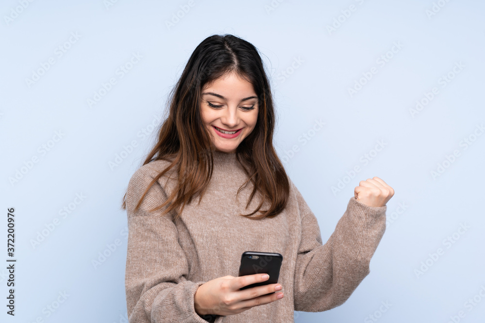 Young brunette woman wearing a sweater over isolated blue background with phone in victory position Young brunette woman wearing a sweater over isolated blue background with phone in victory position