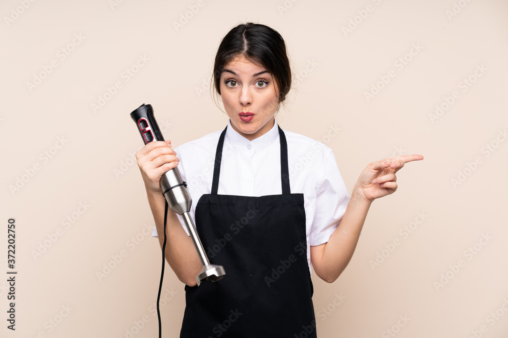 Young woman using hand blender over isolated background surprised and pointing side Young woman using hand blender over isolated background surprised and pointing side
