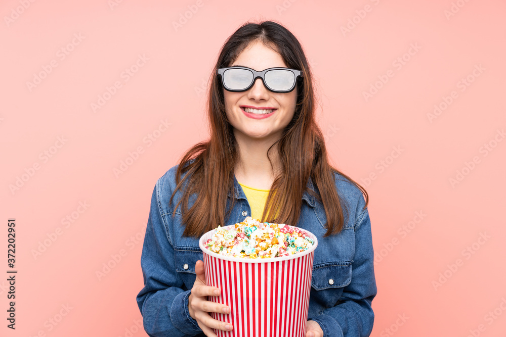 Young brunette woman over isolated pink background with 3d glasses and holding a big bucket of popcorns