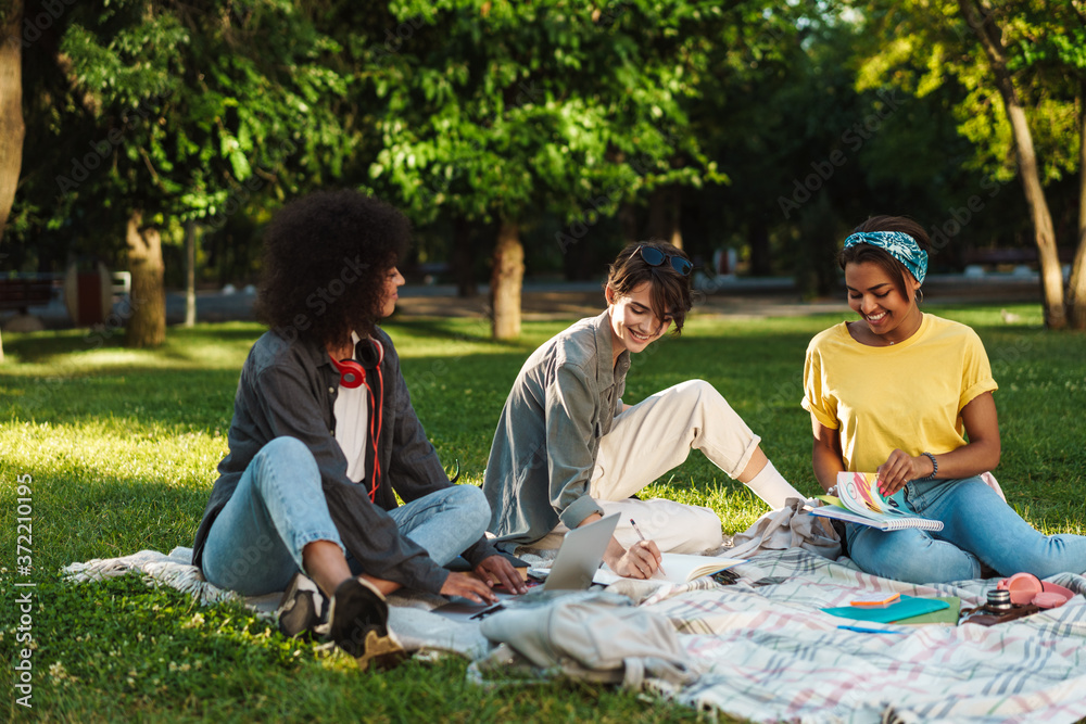 Image of multinational women doing homework with laptop in summer park Image of multinational women doing homework with laptop in summer park