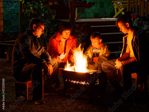 Group of young friends sitting by the fire late at night, grilling sausages and having fun