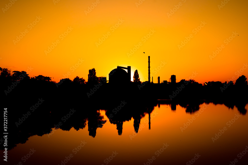 Production facility silo at sunset.  Industrial complex located by the lake. Grain or other loose product storehouse silhouette on the horizon. Water reflection. 