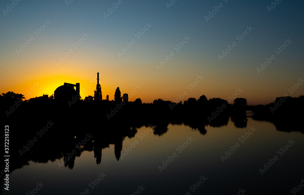 Production facility silo at sunset.  Industrial complex located by the lake. Grain or other loose product storehouse silhouette on the horizon. Water reflection. 