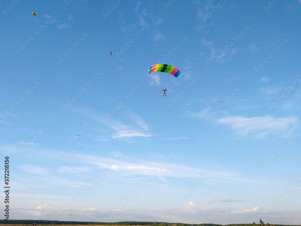 A skydiver with a bright multicolored parachute flies against the background of a blue sky with white sparse clouds A skydiver with a bright multicolored parachute flies against the background of a blue sky with white sparse clouds