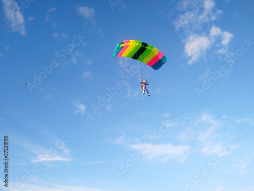 A skydiver with a bright multicolored parachute flies against the background of a blue sky with white sparse clouds