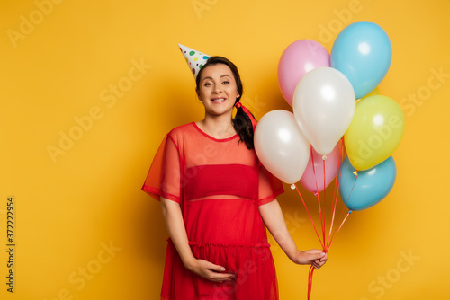 young pregnant woman in party cap holding colorful festive balloons on yellow