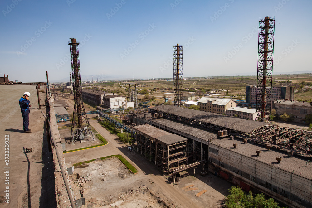 Old unused soviet metallurgy factory buildings and rusted chimneys on the blue sky. Worker in white hardhat (left) on the roof. Taraz city, Kazakhstan.