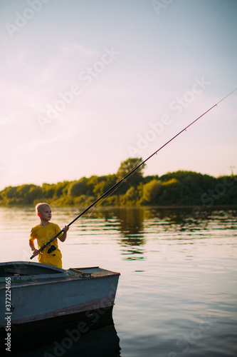 Stylish baby boy fisherman with a fishing rod in hand fishing from boat on river in summertime