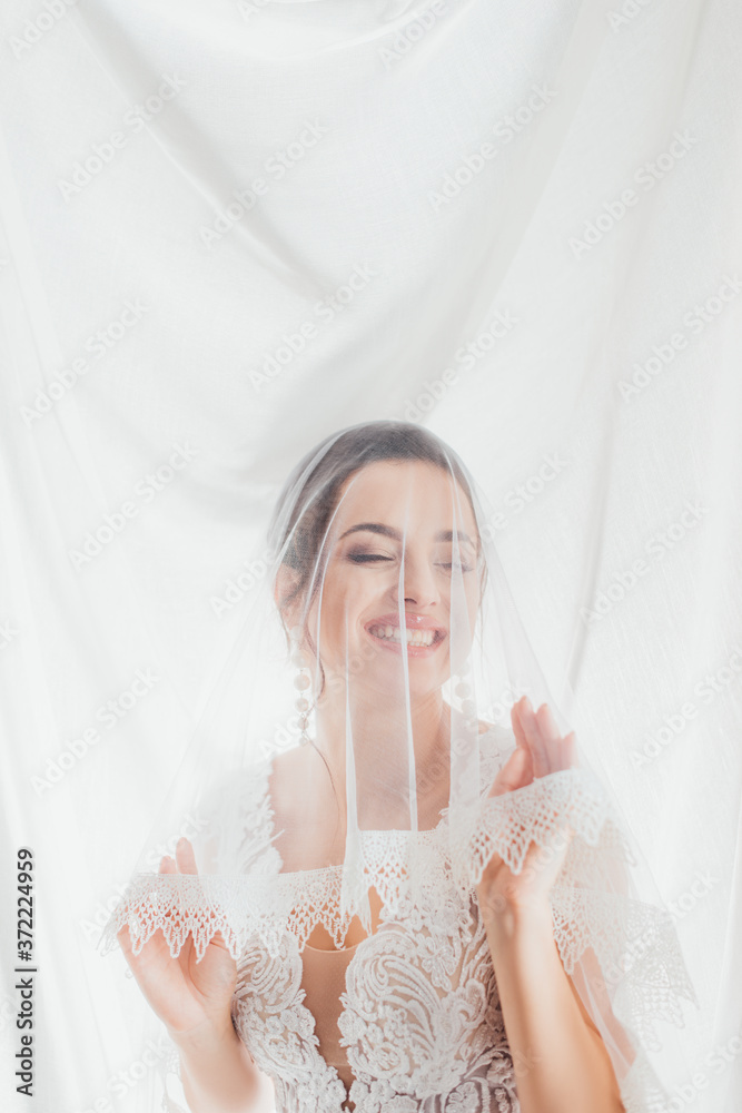 Brunette bride in wedding dress touching lace veil near white cloth Brunette bride in wedding dress touching lace veil near white cloth