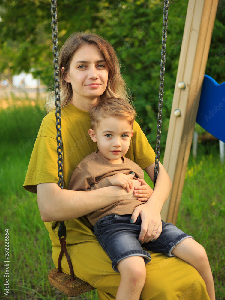 Cute baby boy and mother ride on swing in playground on green lawn. Summertime photo for ad or