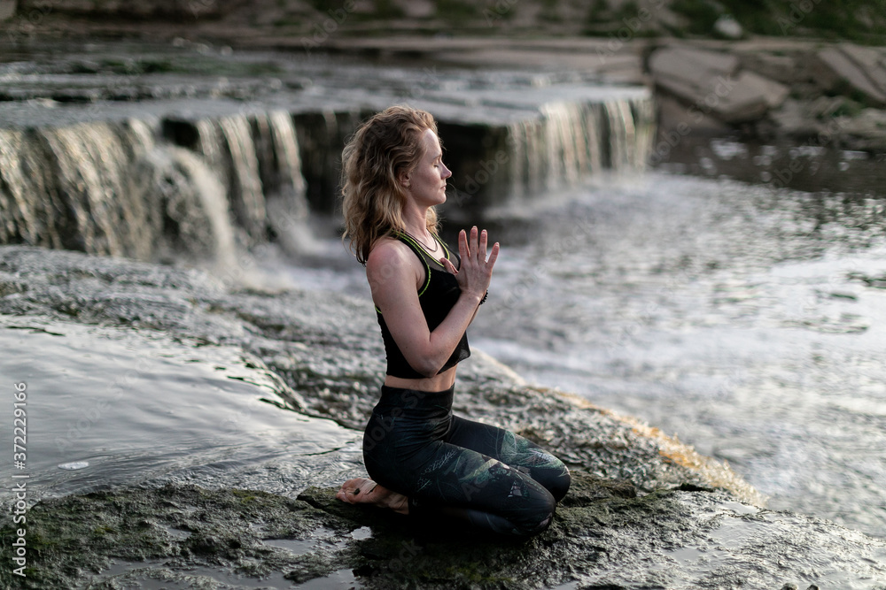 Blonde young woman in sportswear practicing breathing yoga outdoors in harmony with nature. Fitness girl meditating with closed eyes on the edge of waterfall with hands clasped in namaste mudra sign. Blonde young woman in sportswear practicing breathing yoga outdoors in harmony with nature. Fitness girl meditating with closed eyes on the edge of waterfall with hands clasped in namaste mudra sign.