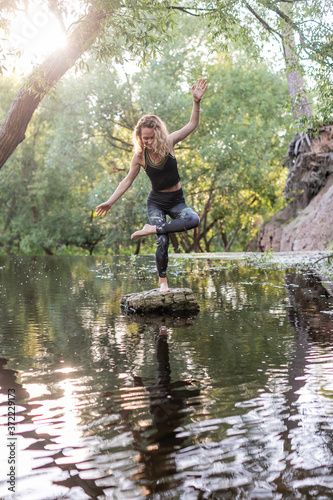 Young woman practicing yoga outdoors in harmony with nature. Fitness girl try to find balance on a small rock in the middle of a forest river © Artem Bruk