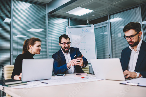 Group of business people working in office with electronic devices, team of experts celebrating victory of achievement of overjoyed male colleague getting good news about project on smartphone