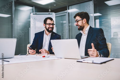 Two office workers in business meeting sitting at desk using computer for business training, professionals smiling and using modern electronics devices during conversation about successful project