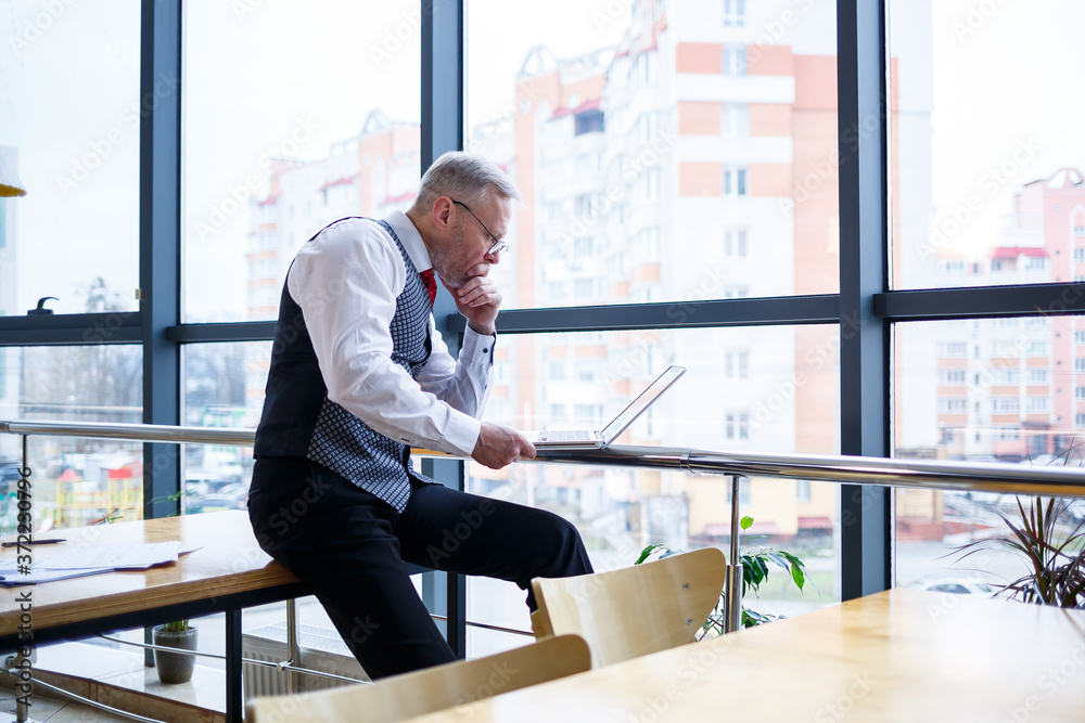 Adult male businessman, teacher, mentor working on a new project. Sits by a large window on the table. He looks at the laptop screen.