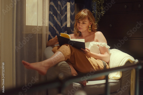 A beautiful girl sits on an armchair in the evening and reads a book. Smart woman, education.