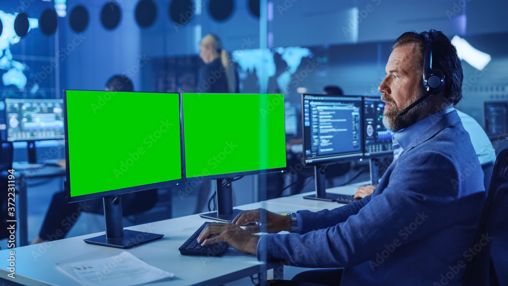 Confident Male Data Scientist Works on Computer with Green Screen Mock Up  Template in Big Infrastructure Control and Monitoring Room. Senior Engineer  in a Call Center Office Room with Colleagues. Stock-Foto