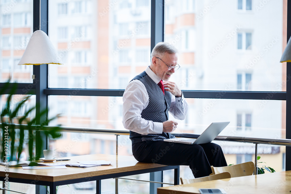 Adult male businessman, teacher, mentor working on a new project. Sits by a large window on the table. He looks at the laptop screen. Adult male businessman, teacher, mentor working on a new project. Sits by a large window on the table. He looks at the laptop screen.