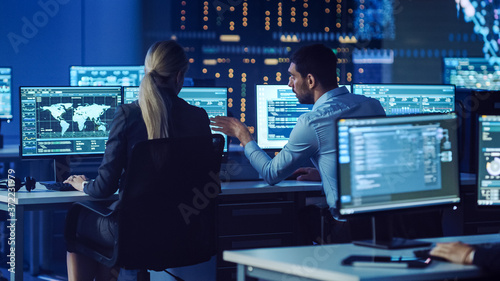 Confident Female Data Scientist Works Closely to her Young Colleague in Big Control and Monitoring Room with Neural Network. Young Specialists Talk and Discuss Work in a Room with Other Employees. © Gorodenkoff