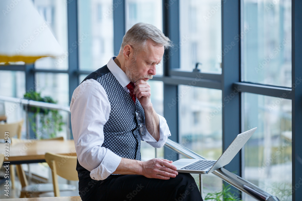 Adult male businessman, teacher, mentor working on a new project. Sits by a large window on the table. He looks at the laptop screen. Adult male businessman, teacher, mentor working on a new project. Sits by a large window on the table. He looks at the laptop screen.