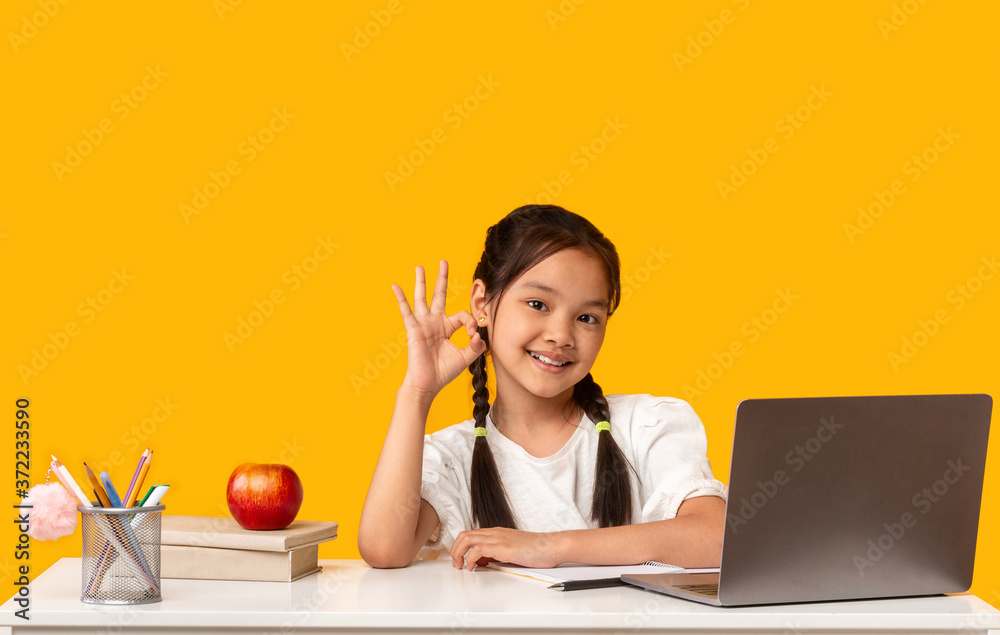 Korean School Girl Gesturing Okay Sitting At Desk, Yellow Background Korean School Girl Gesturing Okay Sitting At Desk, Yellow Background