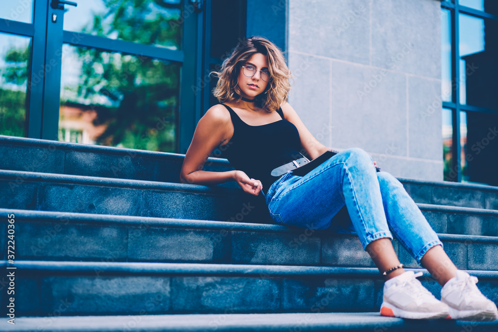 Portrait of serious female hipster in stylish eyewear sitting on stairs during free time outdoors, gorgeous girl in spectacles and fashionable clothes looking at camera lying on payment stairs Portrait of serious female hipster in stylish eyewear sitting on stairs during free time outdoors, gorgeous girl in spectacles and fashionable clothes looking at camera lying on payment stairs