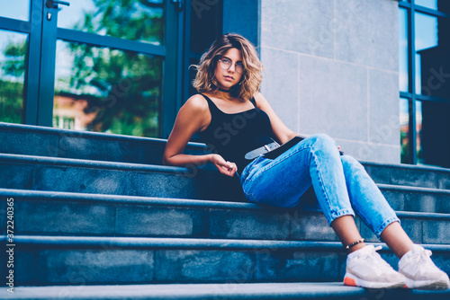 Portrait of serious female hipster in stylish eyewear sitting on stairs during free time outdoors, gorgeous girl in spectacles and fashionable clothes looking at camera lying on payment stairs