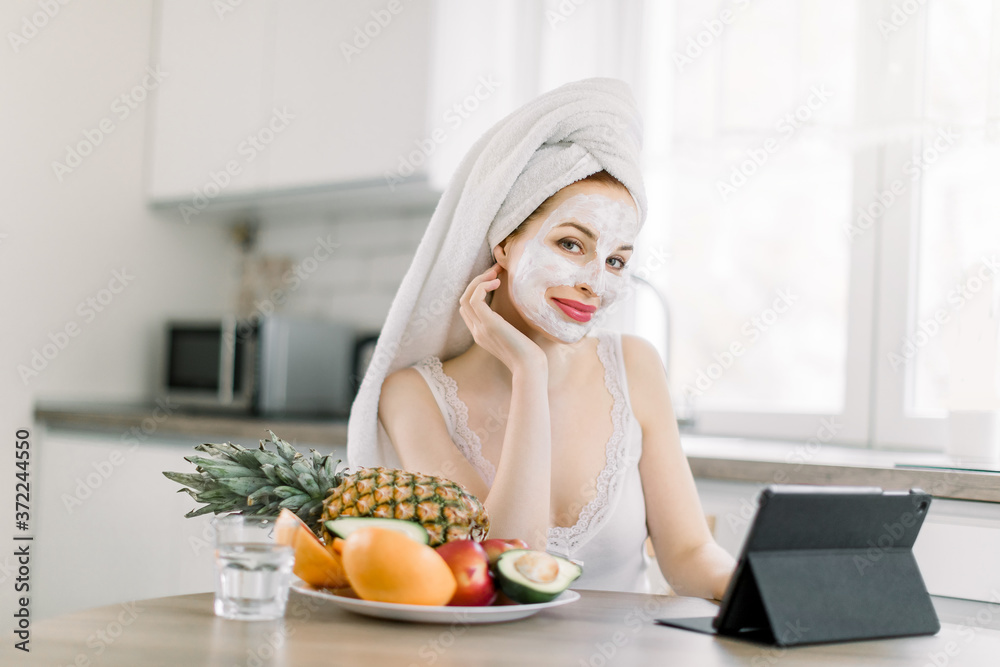 Smiling Caucasian young woman with white anti aging facial mask on face, studying, working or doing online shopping on tablet computer, while sitting at the kitchen table with fruits at home Smiling Caucasian young woman with white anti aging facial mask on face, studying, working or doing online shopping on tablet computer, while sitting at the kitchen table with fruits at home
