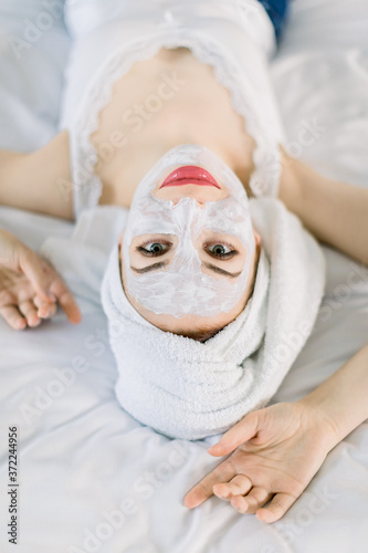 Top close up view of charming smiling Caucasian woman with white clay facial mask and towel on her head, lying on bed and looking at camera, while enjoying skin care treatment
