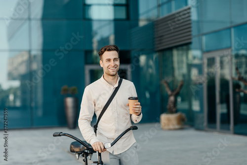 Coffee takeaway and eco transport. Smiling man holding glass and go with bicycle near office building
