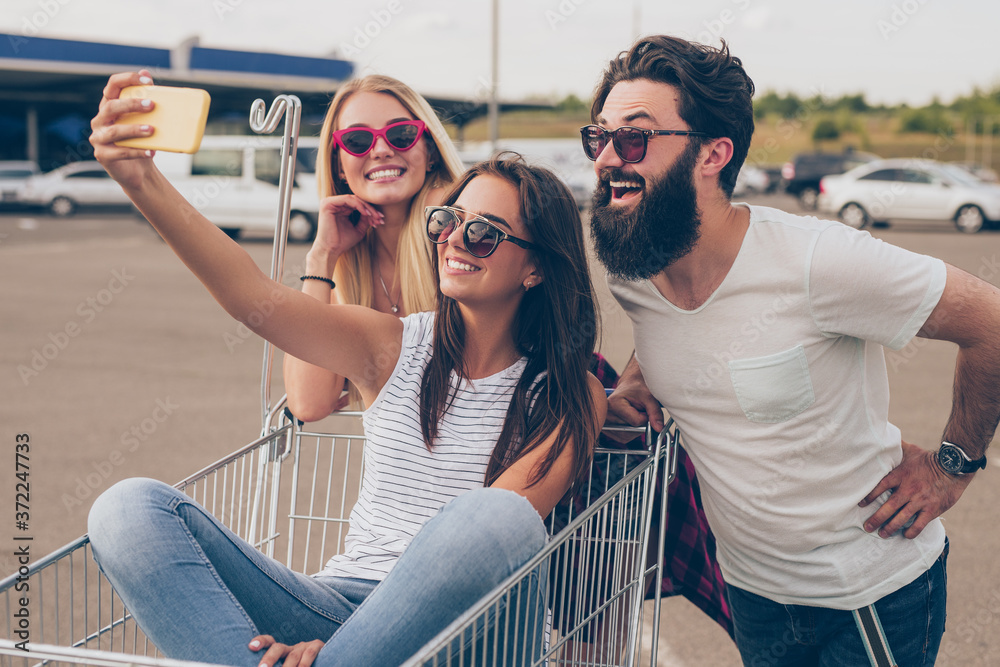 Cheerful young friends with shopping cart taking selfie on street