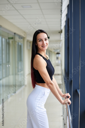 Young brunette woman, wearing white pants and black top, standing in light passageway, posing, leaning on railing by the window. Businesswoman on a break. Female portrait photography.