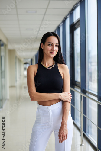Young brunette woman, wearing white pants and black top, standing in light passageway with huge windows, posing. Businesswoman on a break. Female portrait photography.