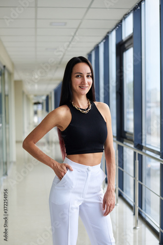 Young brunette woman, wearing white pants and black top, standing in light passageway with huge windows, posing. Businesswoman on a break. Female portrait photography. Free Download Young brunette woman, wearing white pants and black top, standing in light passageway with huge windows, posing. Businesswoman on a break. Female portrait photography. - GFXLoad