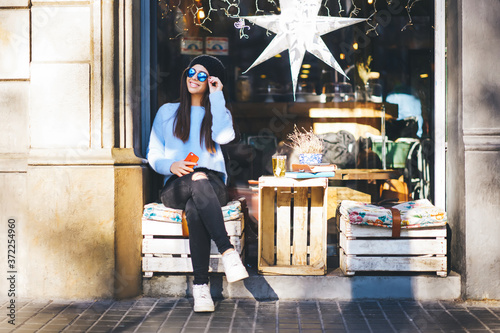 Foto Dreamy smiling female traveller in stylish wear and trendy sunglasses sitting in cafe and enjoying good weather in new town